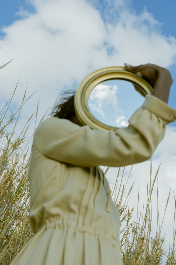 Artistic portrayal of a woman holding a mirror reflecting the sky in a field, emphasizing fashion and surrealism.
